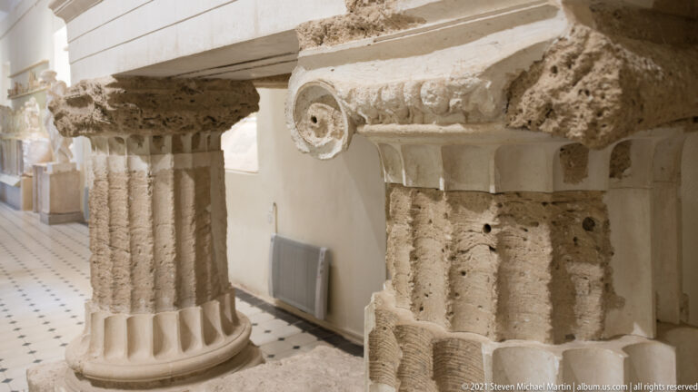 Two reconstructed fluted stone columns with weathered carved capitals, including a prominent spiral volute, in a museum gallery.