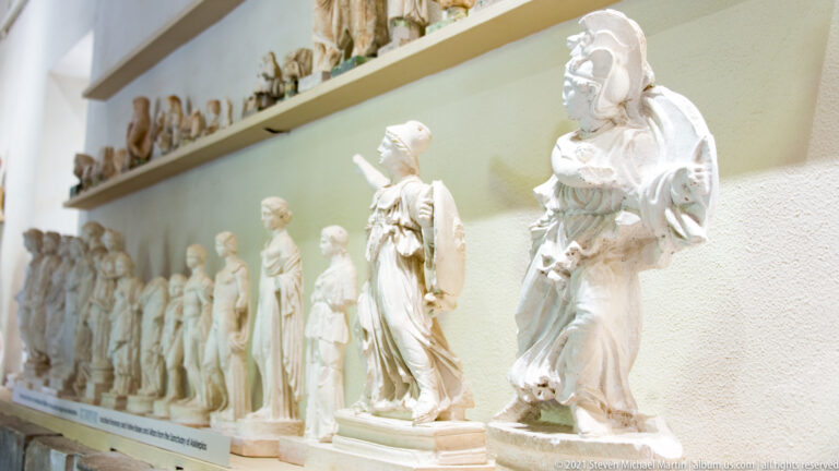 Row of small classical figurative sculptures on a museum shelf, with a larger draped female figure in the foreground.