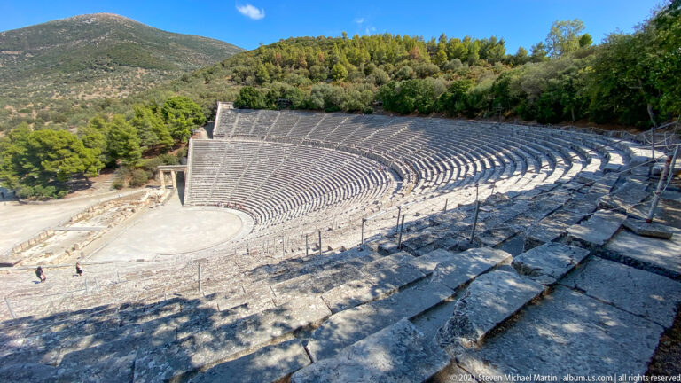 High oblique view across weathered stone seating toward the circular orchestra and opposite cavea of the ancient theatre.