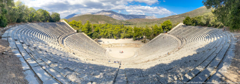 Very high view down the theatre’s steep stone seating toward the small orchestra, trees, and mountains beyond.