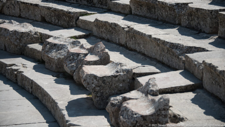 Close detail of worn stone theatre benches and projecting broken seat ends casting sharp shadows.