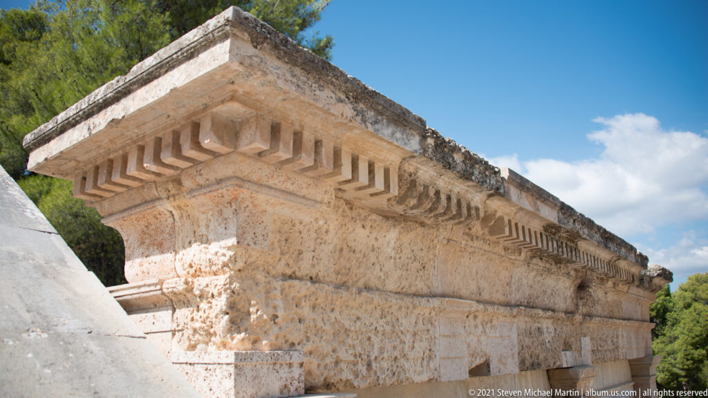 Upward view of massive pale ashlar masonry and a projecting block cornice beside the theatre seating.