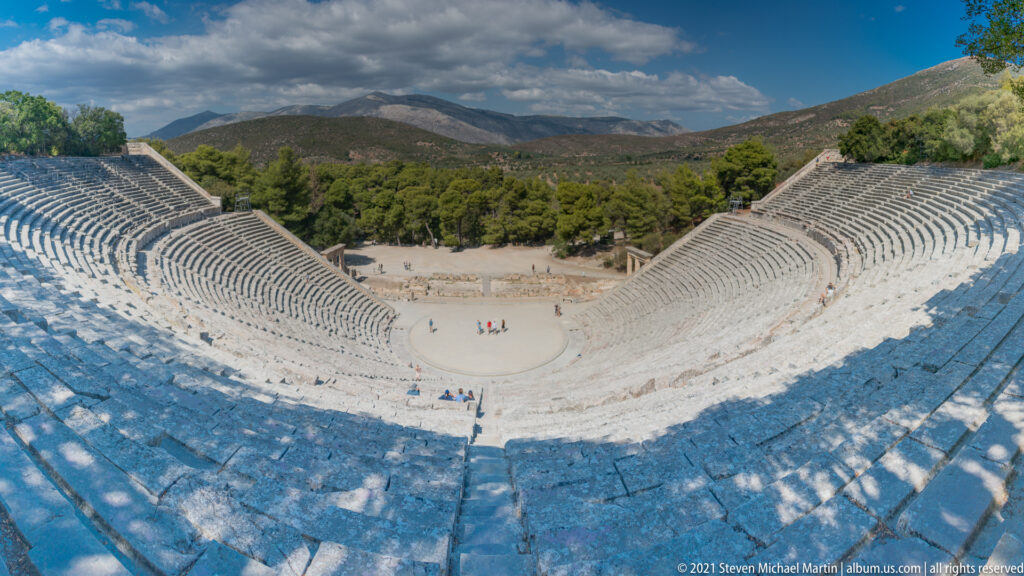 Wide upper view of the full stone cavea and orchestra with green trees and mountain ridges beyond.
