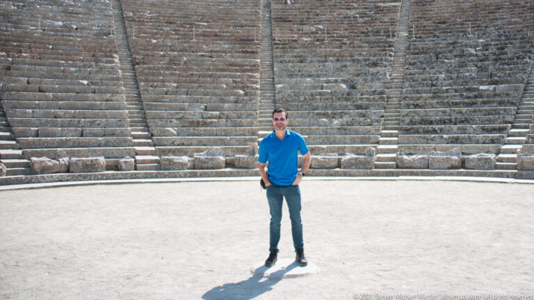 Man in a blue shirt standing on the pale orchestra floor with steep stone theatre seating rising behind him.