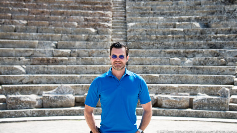 Waist-up portrait of a smiling man in a blue shirt and sunglasses before blurred ancient stone seating.