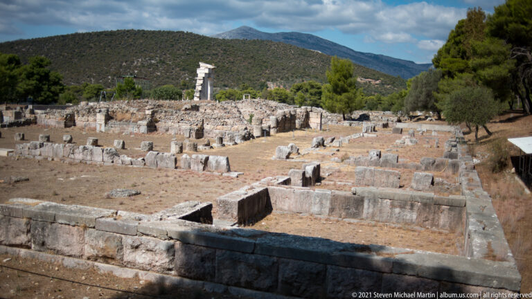Low stone foundations and scattered blocks across the Asklepieion ruins, with one reconstructed vertical element and hills beyond.