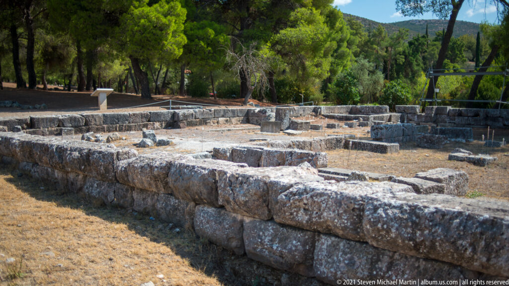 Low rectangular stone walls and room foundations of an ancient bath complex among dry grass and pine trees.