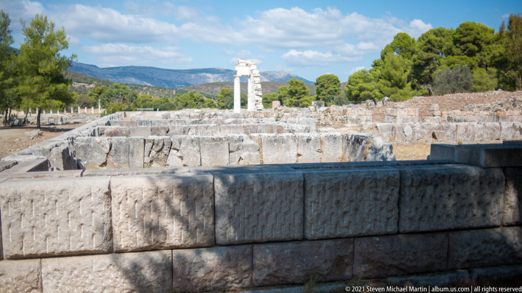 Large stone foundation blocks in the foreground with a reconstructed upright element among sanctuary ruins and pine-covered hills.
