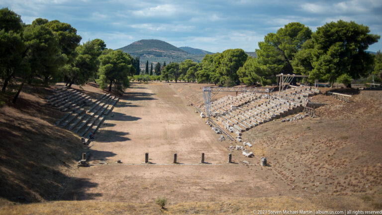 Long dry ancient stadium track with stone spectator seating along both sides and a wooded mountain in the distance.