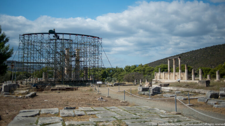 Large steel scaffolding around a reconstructed monument beside a row of standing columns and ancient stone foundations.