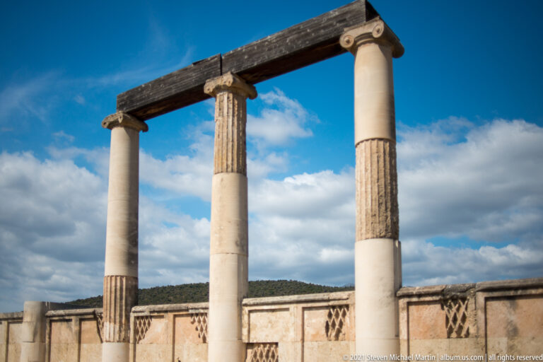 Three reconstructed classical columns with mixed smooth and fluted drums supporting a short dark entablature against blue sky.