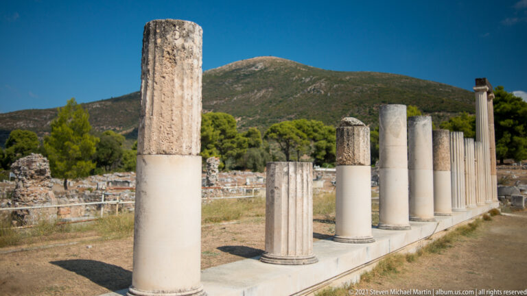 Diagonal row of reconstructed and fragmentary stone columns with fluted sections and pale replacement drums against a mountain.