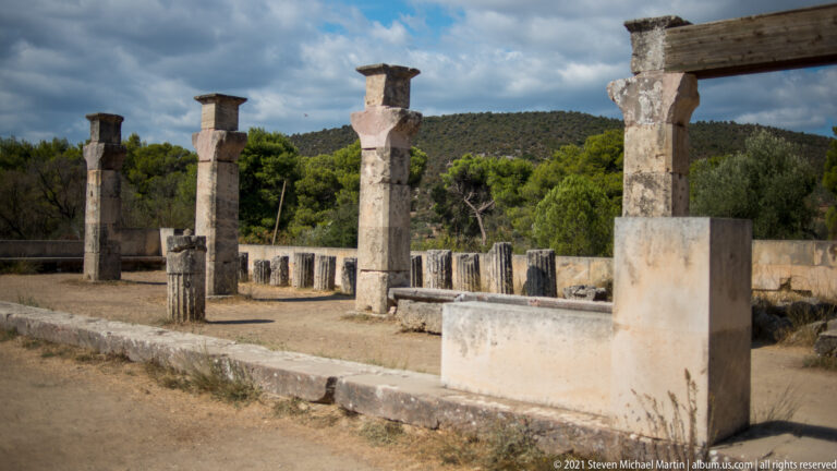 Partial row of reconstructed stone supports with short beam sections on a low platform amid scattered architectural fragments.