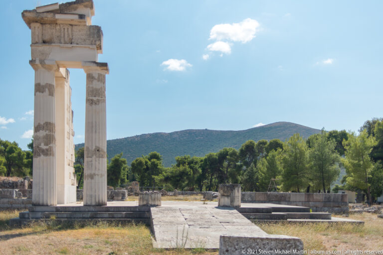 Two tall reconstructed fluted columns supporting a short entablature above low ruins with green hills behind.