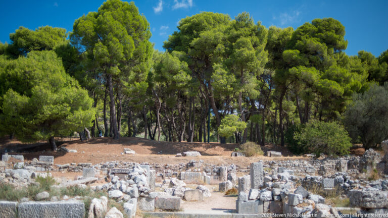 Low stone foundations and scattered blocks spread across a wooded archaeological area beneath tall pine trees.