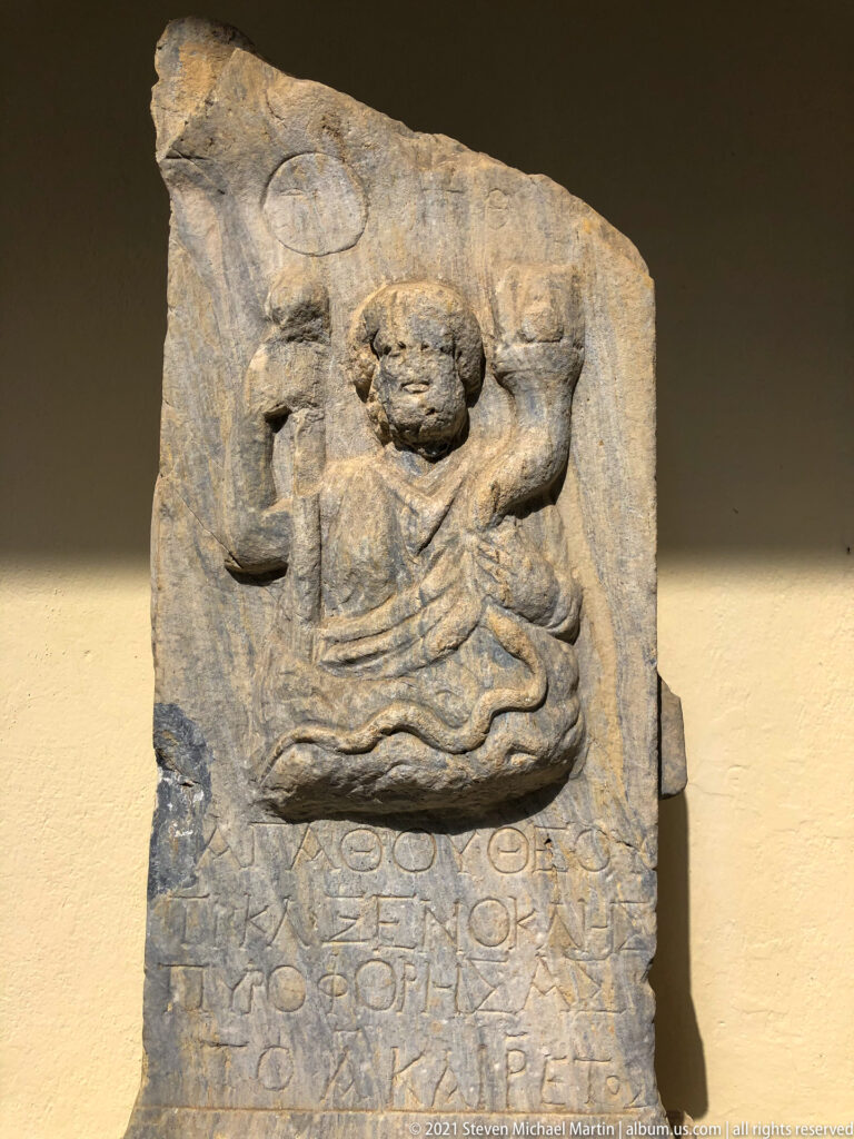 Weathered stone relief of a male figure above several lines of carved Greek inscription.