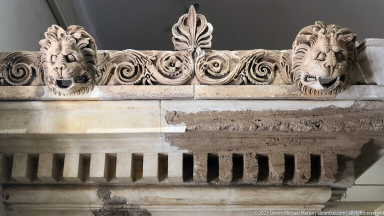 Marble architectural fragment with two lion heads, scrolling foliage, central palmette, and a row of dentils.