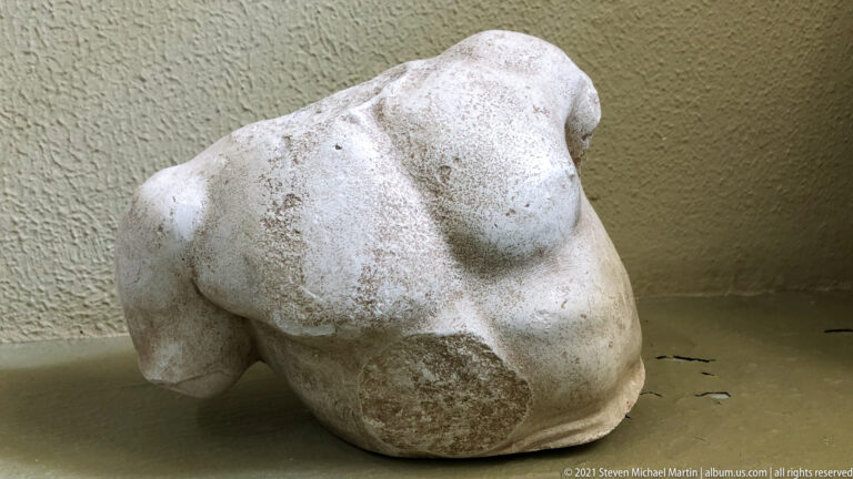 Fragmentary white marble male torso with muscular chest and shoulders, missing head, arms, and lower body.