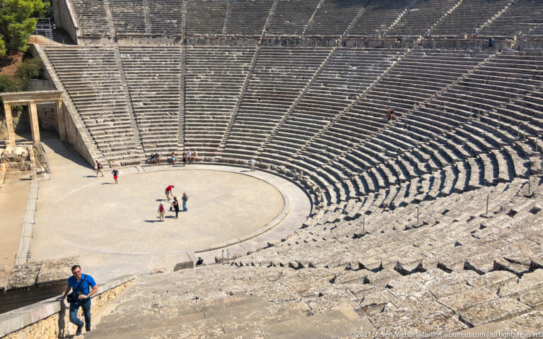 High side view of the circular orchestra and steep curved seating with several visitors inside the Ancient Theatre at Epidaurus.
