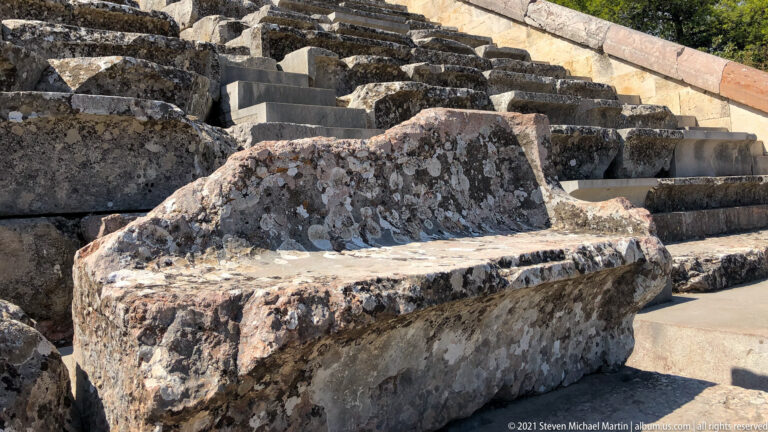 Weathered high-backed stone seat among stepped rows of simpler ancient theatre benches.