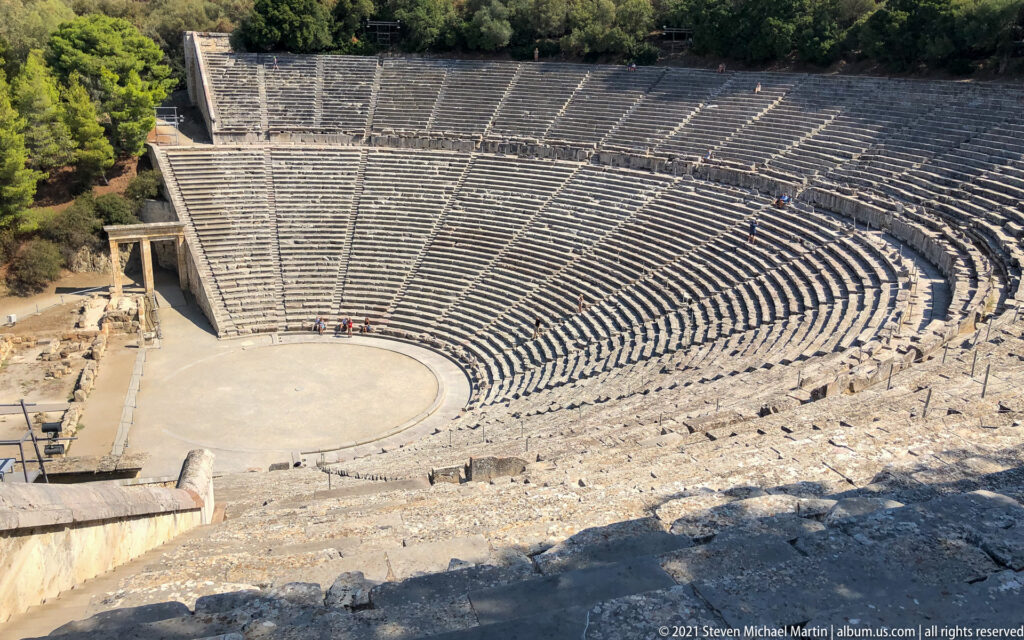 High oblique overview of the circular orchestra and sweeping stone cavea bordered by trees.