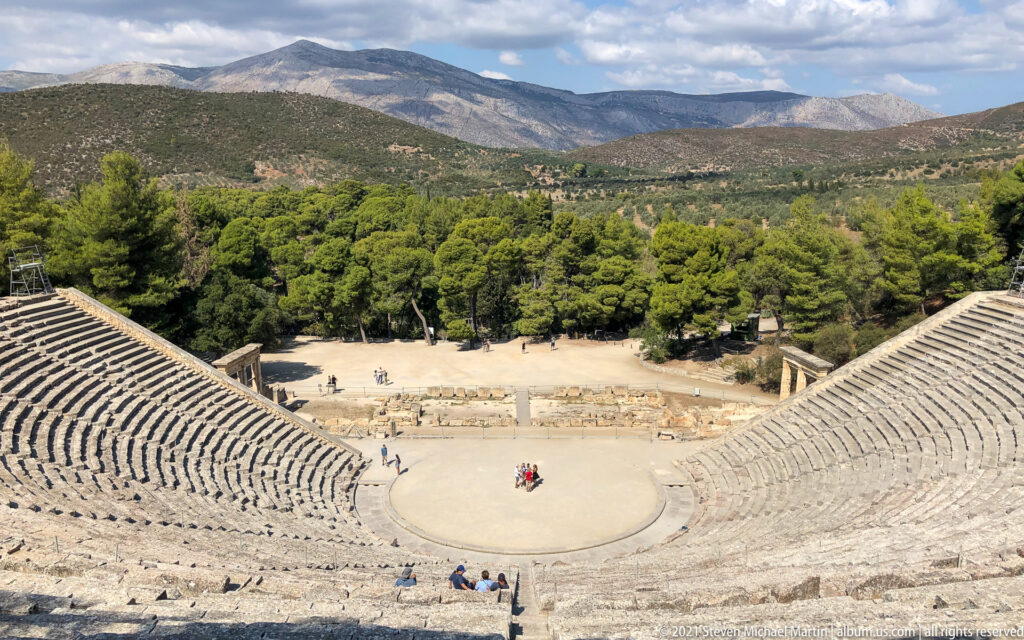 Centered high view down the theatre to the circular orchestra, pine forest, and layered mountains beyond.