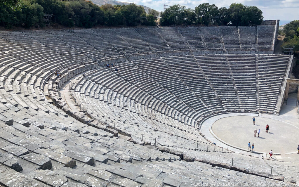 High lateral view over rough foreground benches to the orchestra and densely curved seating across the theatre.