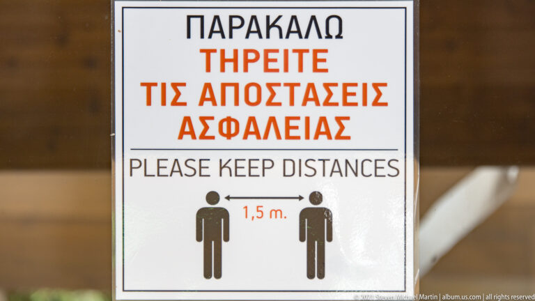 Greek-and-English sign reading “Please keep distances,” with two figures separated by a 1.5-meter arrow.