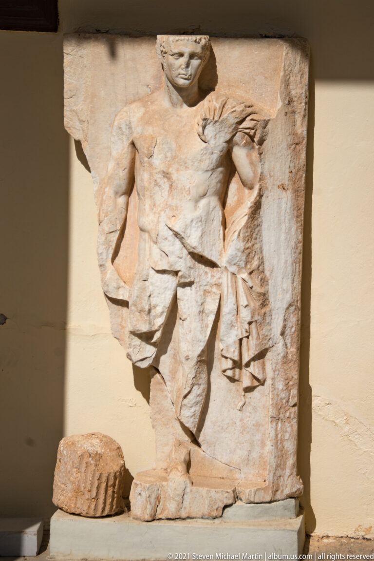 Fragmentary stone relief of a standing male figure in a short draped garment, displayed with smaller carved fragments.