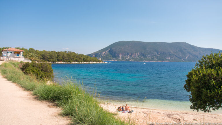 Small pale beach beside turquoise water, with grasses, shrubs, and a steep wooded ridge across the channel.