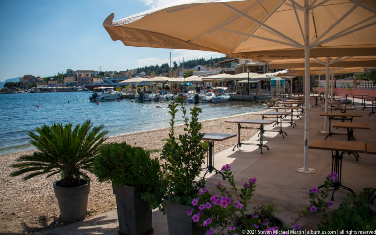 Empty restaurant tables beneath cream umbrellas beside a pebble shore, potted plants, and small boats on the water.