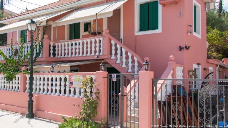 Salmon-pink house with green shutters, white balustrades, exterior stairs, striped awning, and a gated entrance.