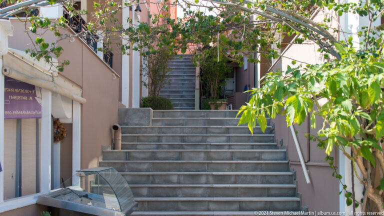 Gray stone steps rising between pastel buildings, with vines overhead and a shopfront at the lower left.