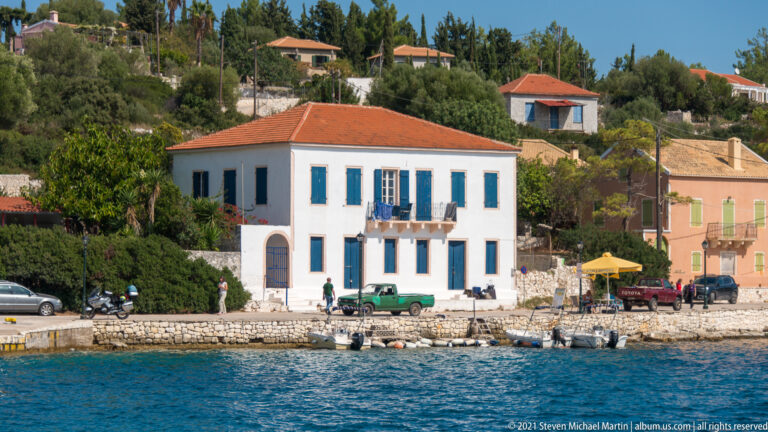 White two-story waterfront house with blue shutters and central balcony, beside a stone quay with small boats.