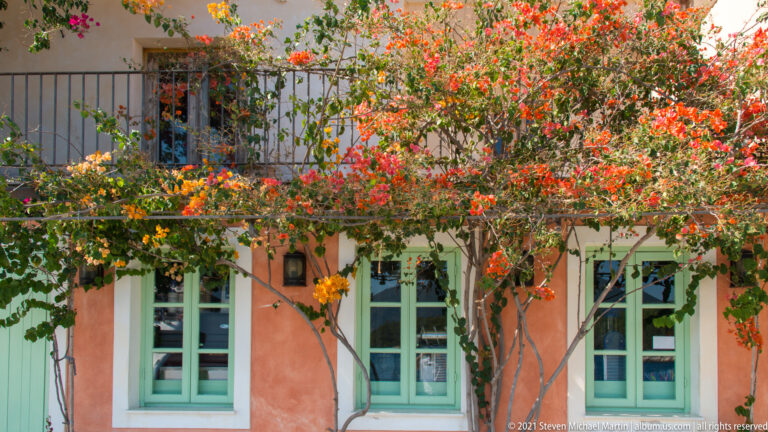 Red and orange bougainvillea covering a salmon wall around three tall pale-green windows.