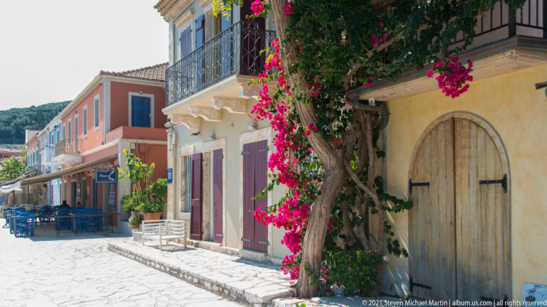 Stone-paved lane beside an arched wooden door and flowering bougainvillea, with pastel buildings and café seating beyond.