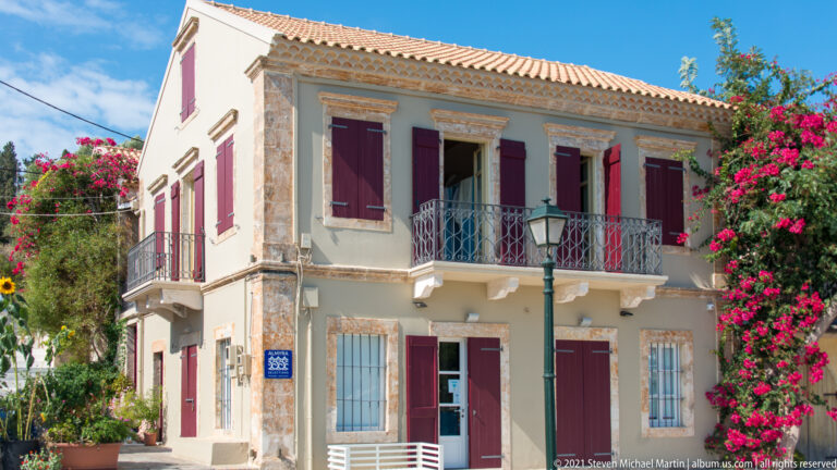Two-story corner house with burgundy shutters, iron balcony, stone trim, red-tiled roof, and bougainvillea at the side.