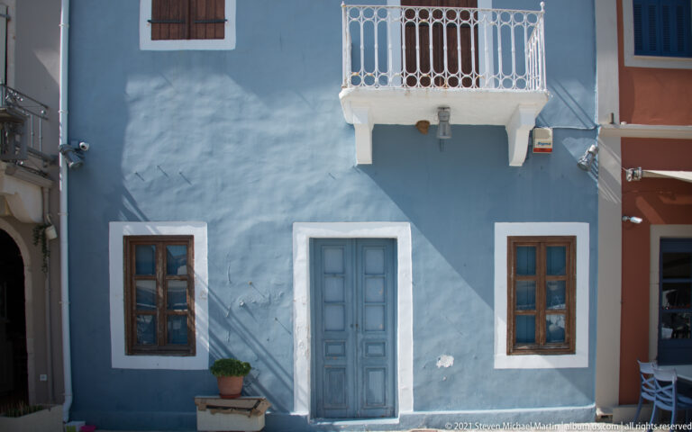 Blue façade with central paneled door, two windows, white trim, and a small white iron balcony overhead.