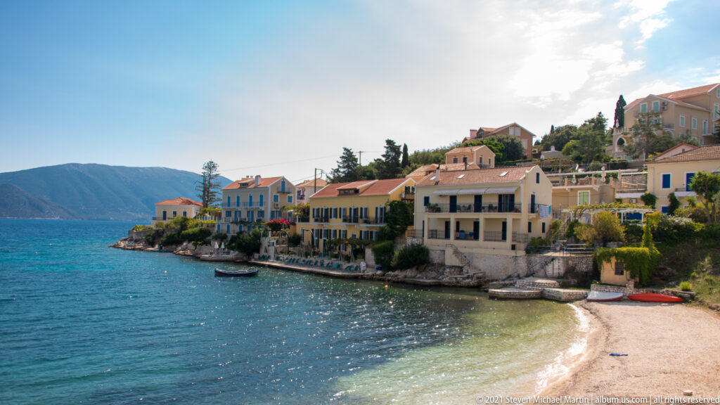 Pastel waterfront buildings with red-tiled roofs beside a small beach and calm blue water at Fiskardo.