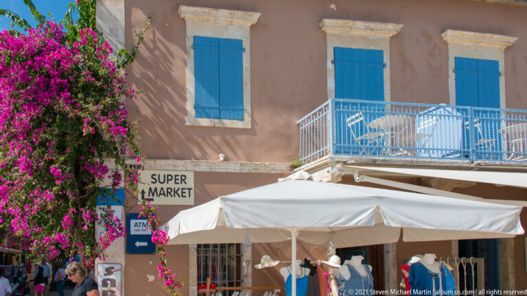 Shopfront under a white awning, framed by magenta bougainvillea, with blue shutters, balcony, supermarket sign, and ATM sign.