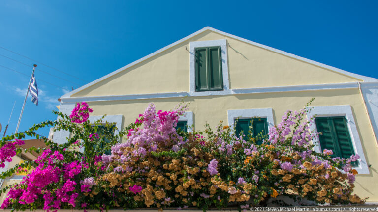 Pale yellow gabled house with green shutters, abundant multicolored bougainvillea, and a Greek flag against blue sky.