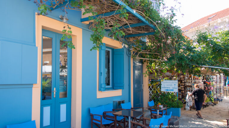 Blue café façade with small tables under a vine-covered pergola, opening onto a narrow lane with shops and a pedestrian.