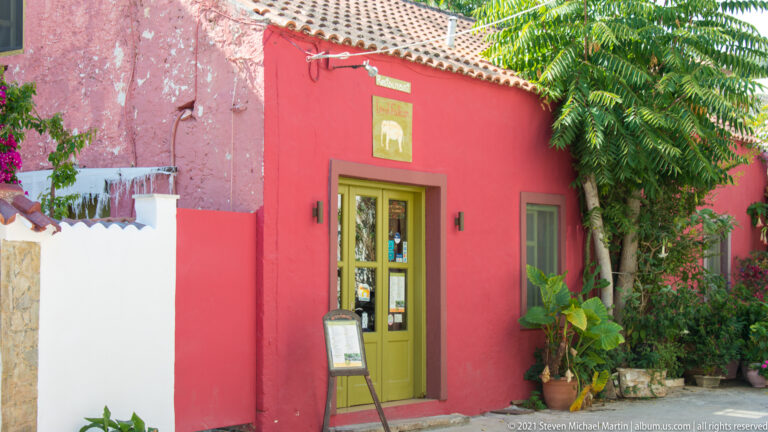 Red-painted building with olive-green door, tiled roof, menu board, potted plants, and dense foliage around the entrance.