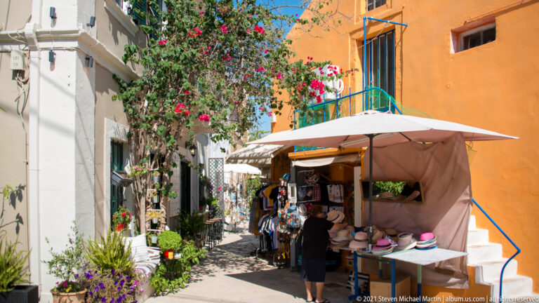 Shopper at a shaded hat and souvenir stall between white and orange buildings on a narrow planted lane.