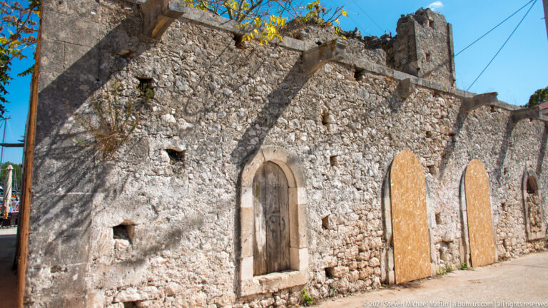 Roofless rough-stone building with tall arched openings, boarded doorways, broken wall tops, and small square holes.