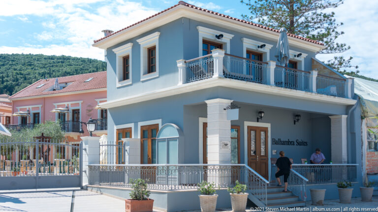Blue-gray Balhambra Suites building with white trim, corner balcony, entrance terrace, potted plants, and hillside behind.