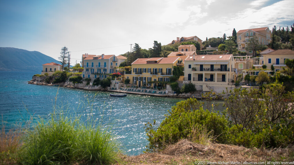 Waterfront buildings and hillside houses at Fiskardo seen across blue water, with grasses and shrubs in the foreground.