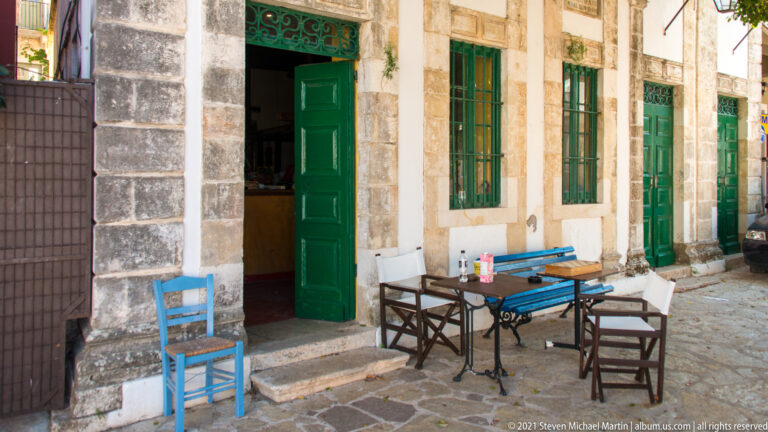 Stone storefront with tall green doors and windows, blue and wooden chairs, and small café tables on the paved lane.