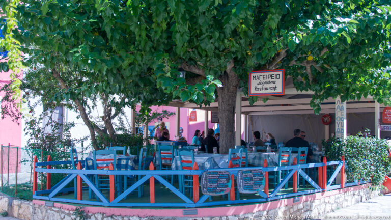 Blue chairs and painted railings surround a shaded restaurant terrace beneath a large tree in Fiskardo.