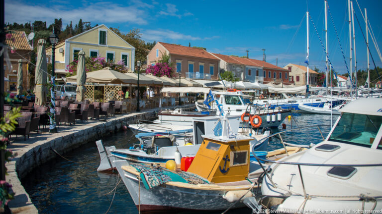 Small yellow-cabin boat moored among sailboats and yachts beside Fiskardo’s restaurant-lined waterfront.
